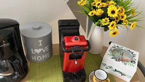 A coffee maker, a trash can, a red espresso machine, a yellow mug, and a vase with yellow flowers on a table.