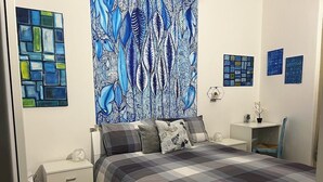 A bedroom with a large bed, a nightstand, a desk, and a chair. There are wall art pieces and a window.