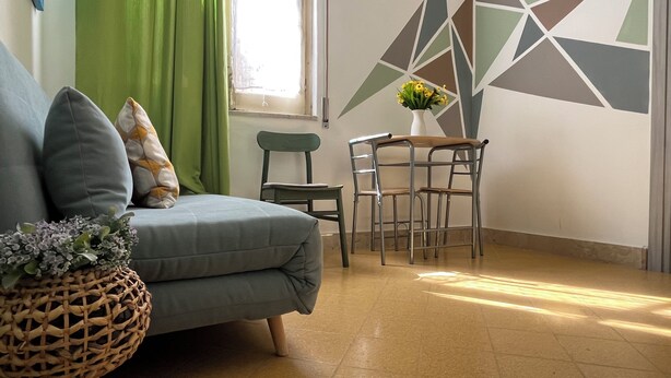 A living room with a sofa, a small table with chairs, a decorative plant, and a geometric wall design.