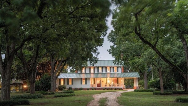 A two-story house with a porch, surrounded by trees and a gravel path.