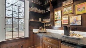 A kitchen with wooden cabinets, a black countertop, and a window with a view of trees. There are shelves with glassware and framed pictures on the wall.