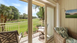 A balcony with a view of a golf course, a wicker chair, and a table. - 519 Plantation Club | Steps to Pool & Short Walk to Beach Access (Hilton Head Island)