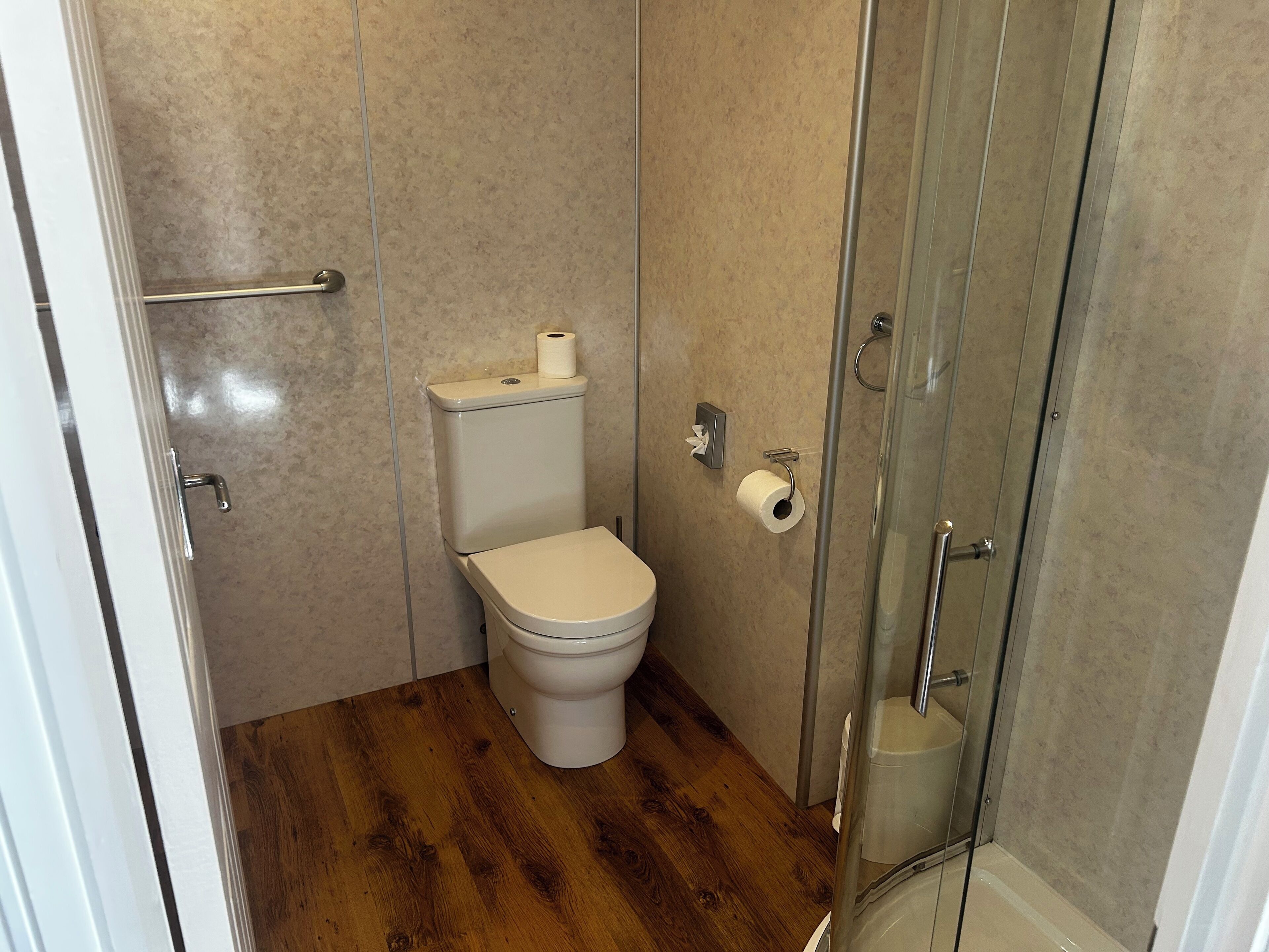 A small, modern bathroom with a toilet, a glass shower enclosure, and a wooden floor.