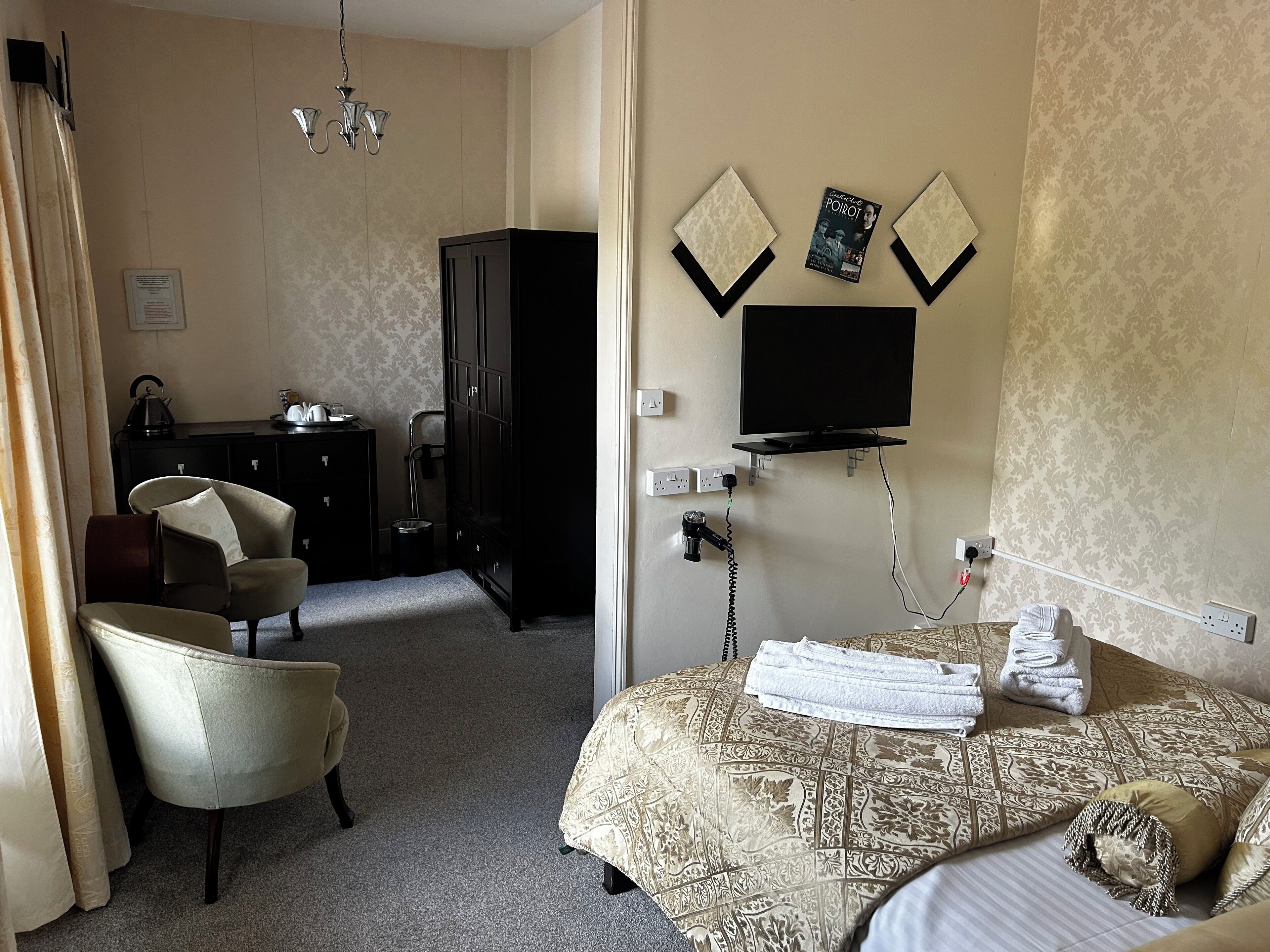 Quarto de hotel com cama, poltrona, escrivaninha, televisão e lustre.