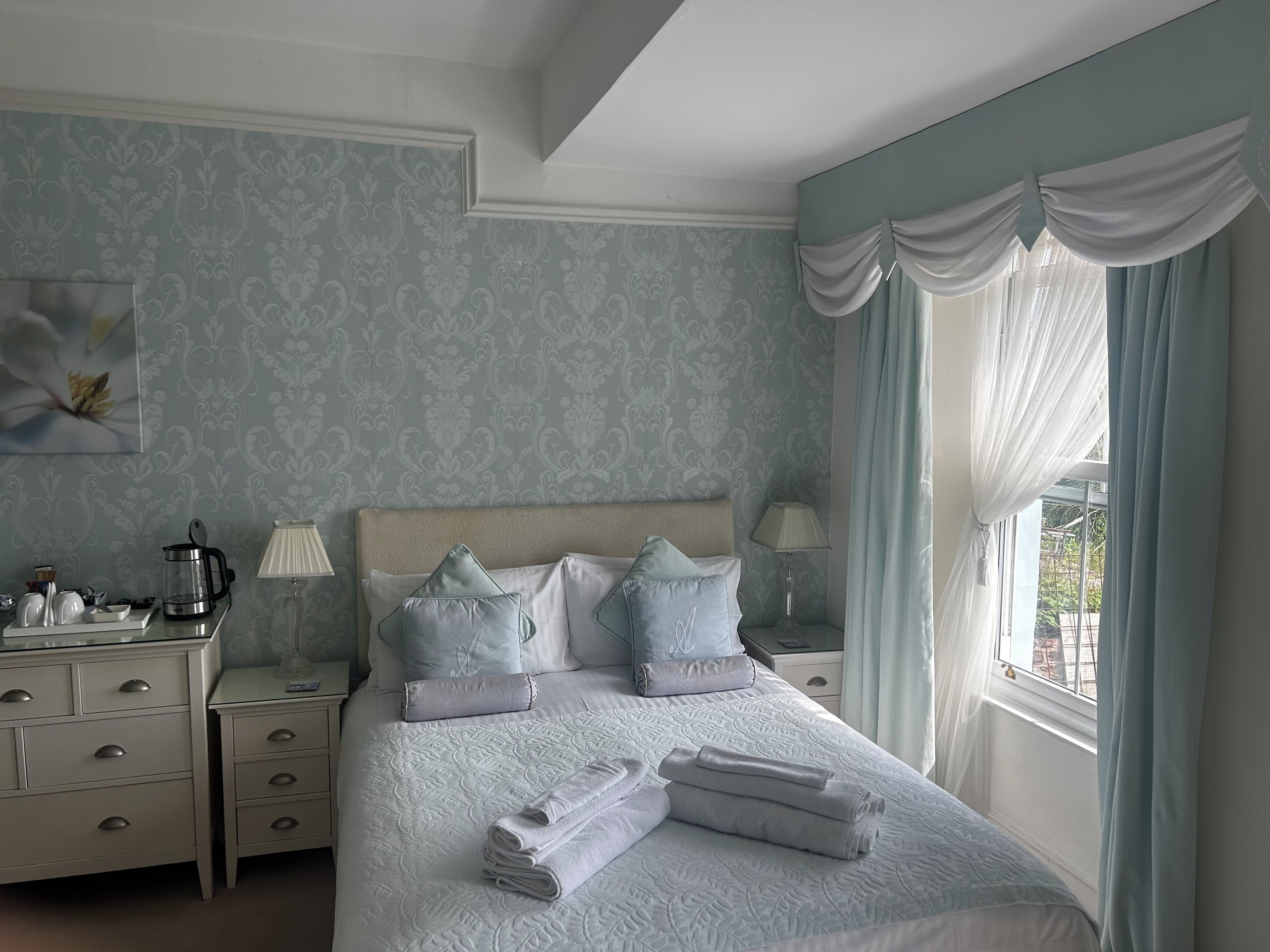 A neatly made bed with pillows, a bedside table with a lamp, and a view of the outdoors through a window with curtains.