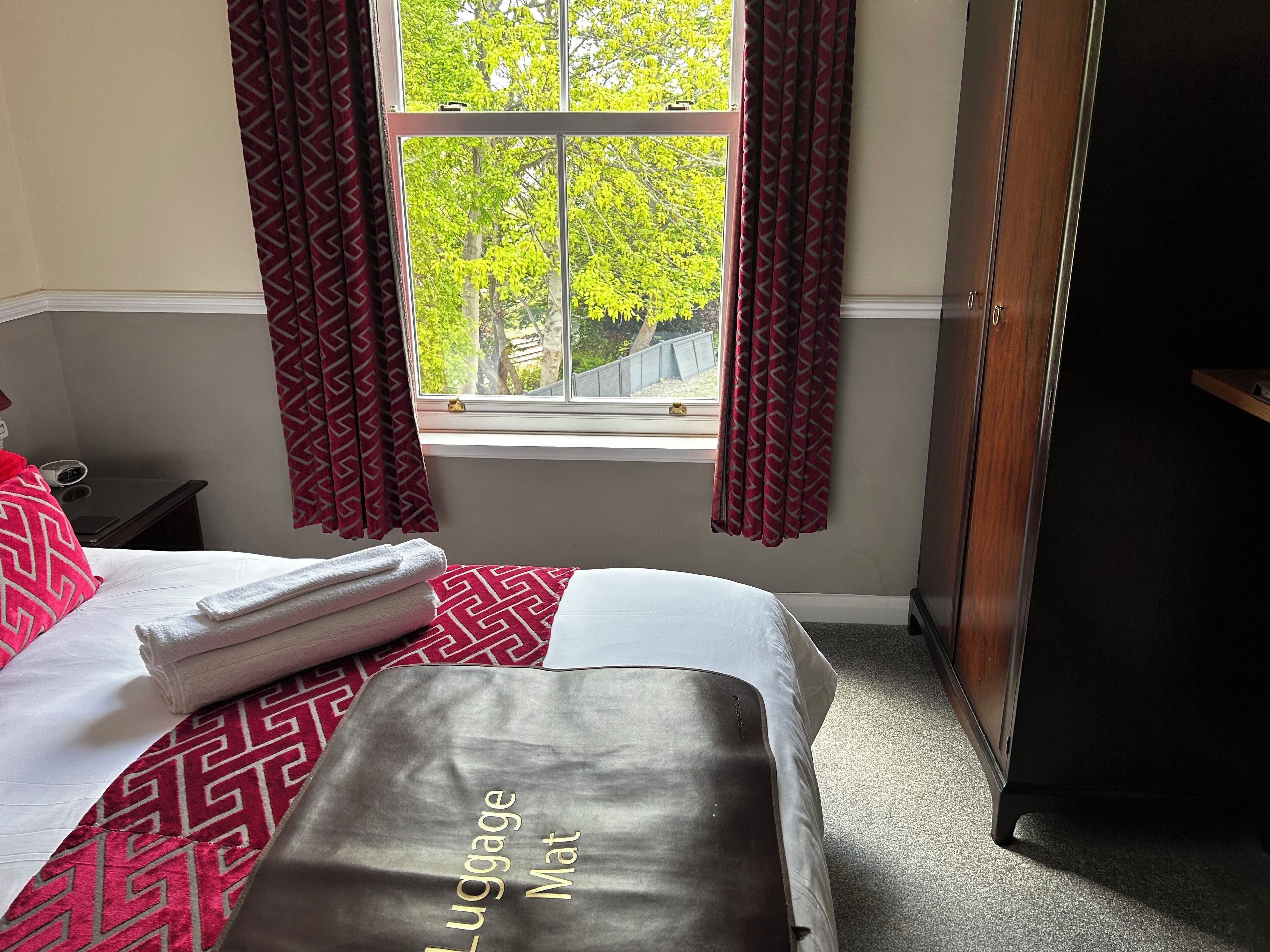 A hotel room with a bed, a suitcase labeled "Luggage Mat," a window with curtains, and a wardrobe.
