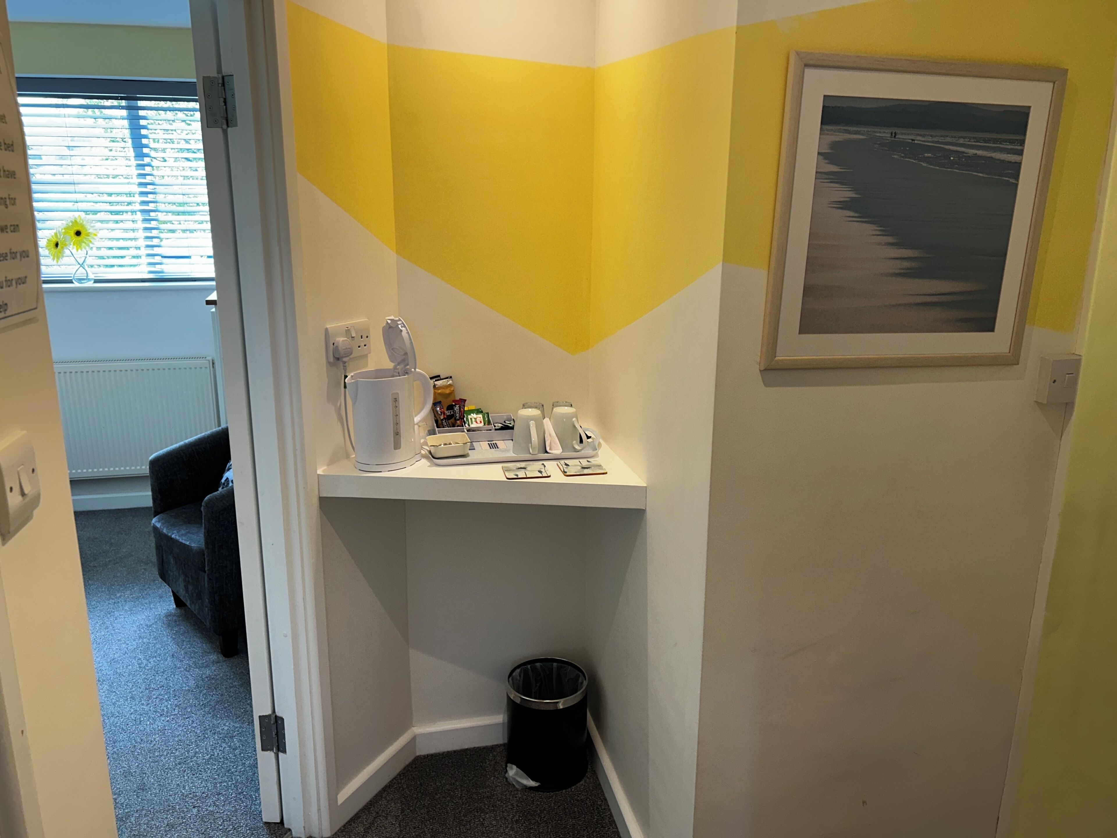 A corner of a room with a white shelf holding a coffee maker, a kettle, and a tray with cups. There's a framed picture on the wall and a black trash bin in the corner.