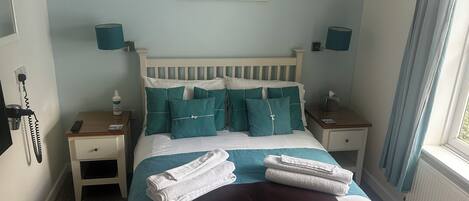 A neatly made bed with teal and white bedding, a luggage mat, and a painting of beach huts on the wall.