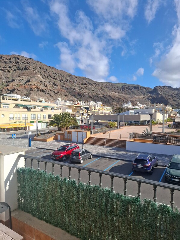 A parking area with several cars, a pedestrian crossing, and a view of a town with buildings and a mountain in the background.