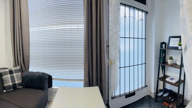 A room with a sofa, a bed, a window with blinds, a door with a metal grid, and a shelf with items. - Brighton charm – Near Beach, City Centre & Station (Brighton and Hove)