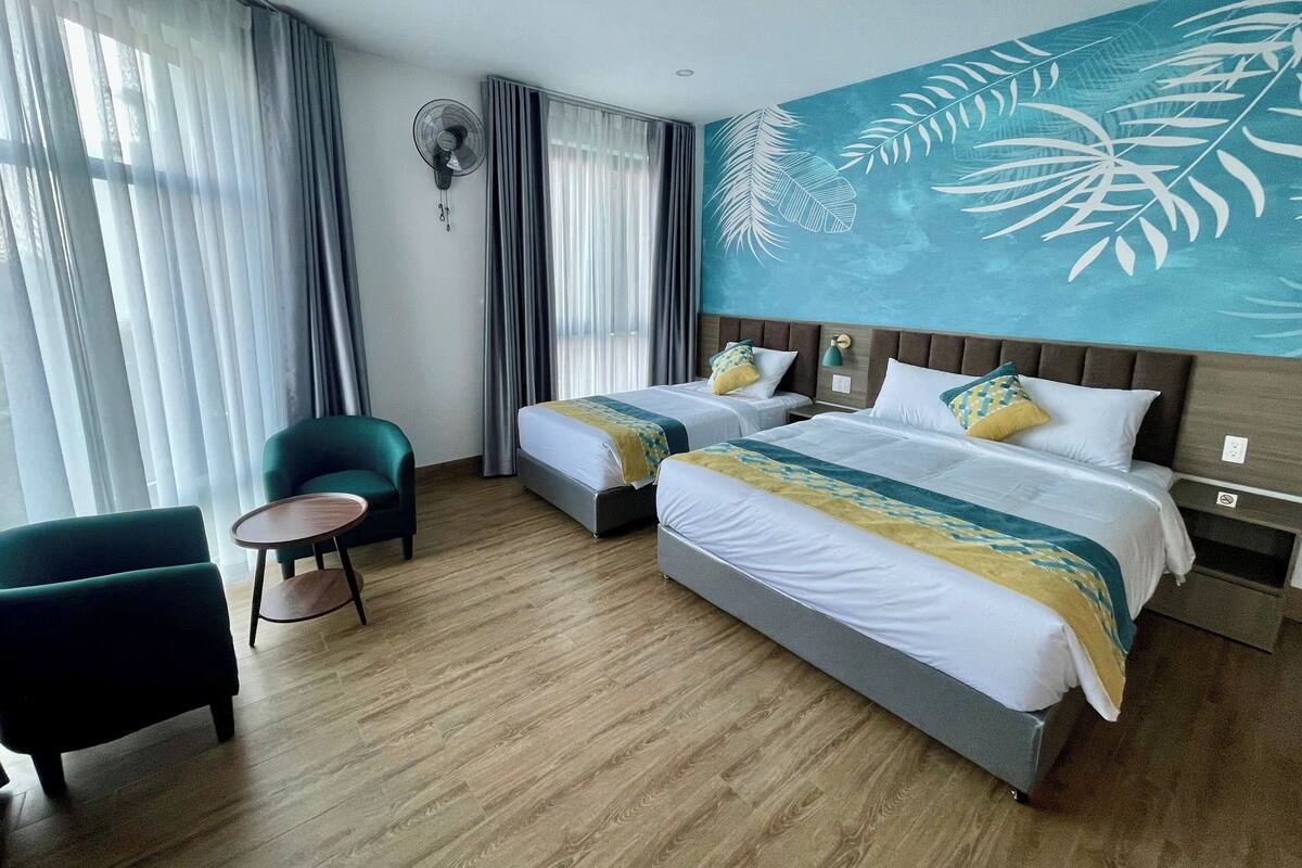 Une chambre d’hôtel avec deux lits, une chaise, une petite table et une grande fenêtre avec des rideaux.