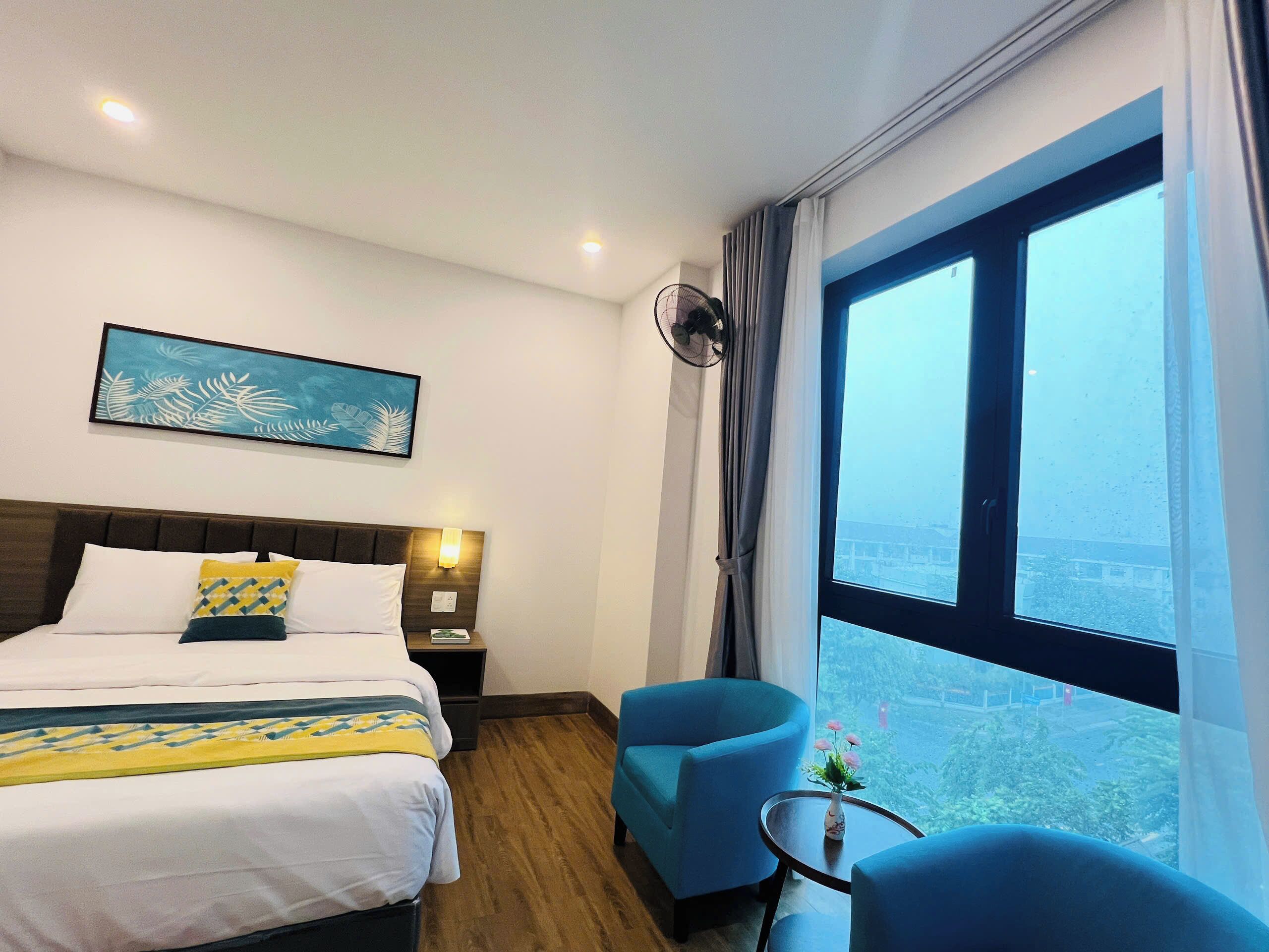 Ein Hotelzimmer mit Bett, Nachttisch, Wandventilator, großem Fenster und Blick auf die Stadt.