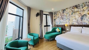 A hotel room with a large bed, two green armchairs, a small table, a wall-mounted fan, and a ceiling light fixture.