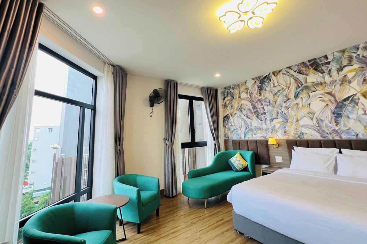 Une chambre d’hôtel avec un grand lit, deux fauteuils verts, une petite table, un ventilateur fixé au mur et un luminaire au plafond.