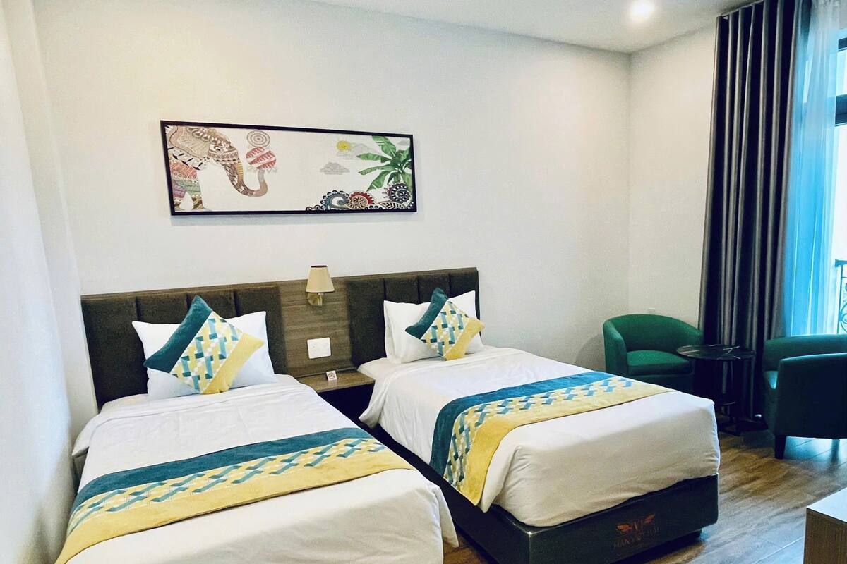 Une chambre d’hôtel avec deux lits, une table de chevet, une lampe, un fauteuil vert et une fenêtre avec des rideaux.