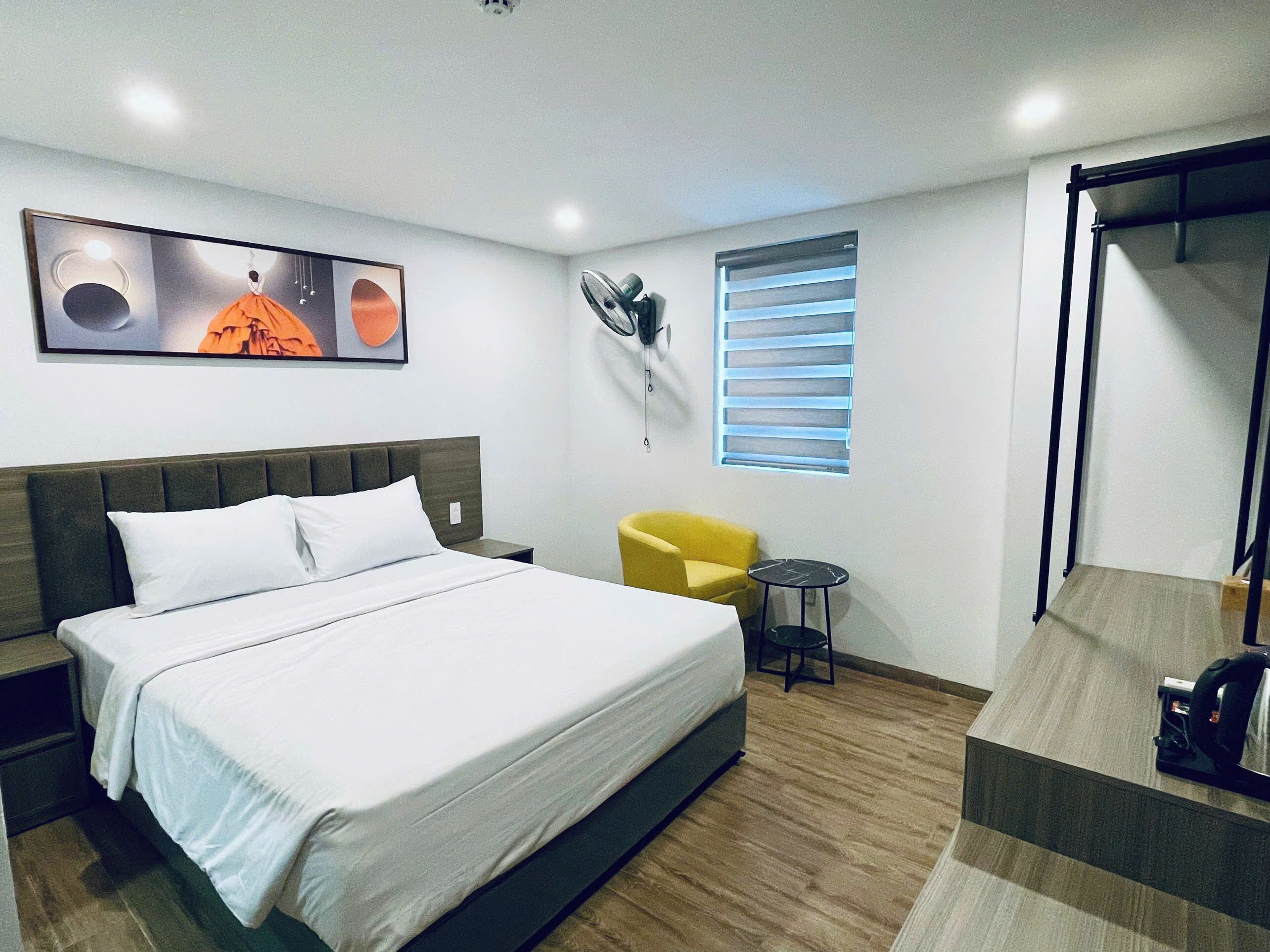 Ein Hotelzimmer mit einem Bett, einem kleinen gelben Sessel, einem Nachttisch, einem Fenster mit Jalousien und einem Deckenventilator.