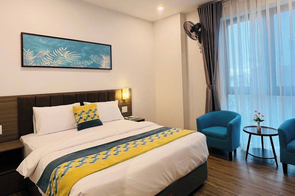 Une chambre d’hôtel avec un lit, deux chaises, une petite table avec un vase, un tableau au mur et une fenêtre avec des rideaux.