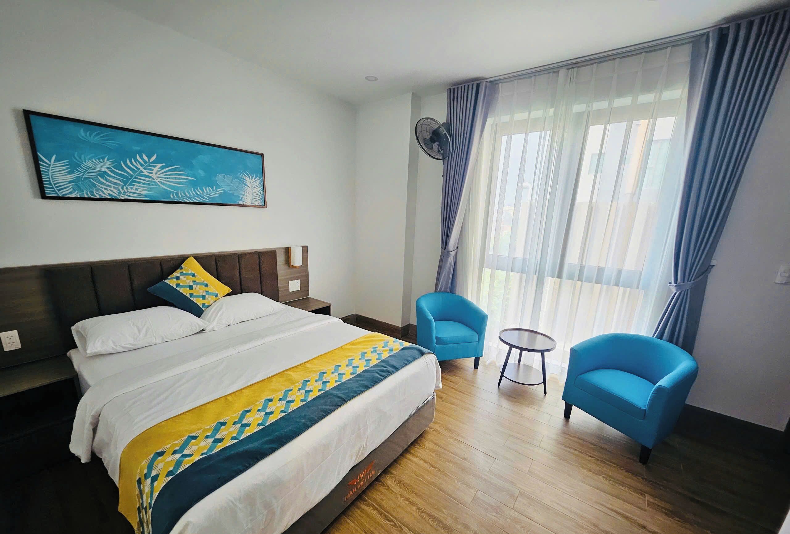 A hotel room with a bed, a blue armchair, a second blue armchair, a small table, and a wall-mounted fan.