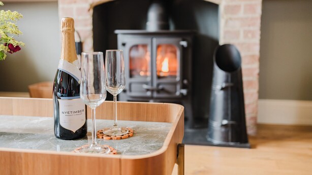 En flaska Nyetimber-vin, två glas och underlägg på ett träbord, med en tegelkamin i bakgrunden.