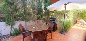 Une terrasse avec une table ronde en bois et des chaises, un barbecue et un parasol.