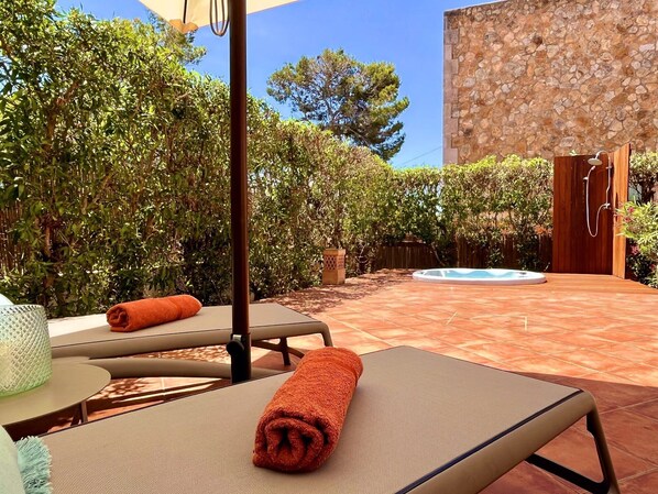A sunlit outdoor area with a hot tub, lounge chairs, and a stone wall.