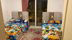 A room with two beds, each with a colorful bedspread featuring cartoon characters. There is a large window with a view of the outside. - Cozy Modern Apartment in El Rehab 2, (Cairo Governorate)
