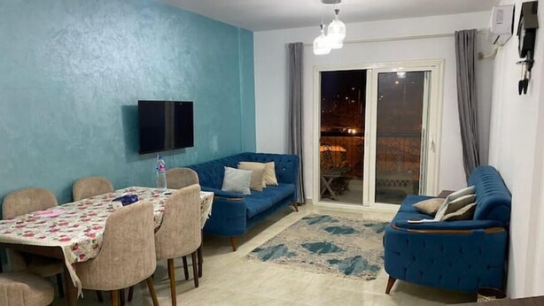 A living room with a blue sofa, a dining table with chairs, and a TV mounted on the wall. - Cozy Modern Apartment in El Rehab 2, (Cairo Governorate)