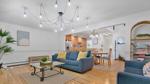 Un salón espacioso con un sofá azul, una mesa de centro y una araña. Se observa un comedor con mobiliario de madera y una cocina al fondo.