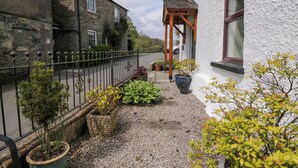Une cour avec un chemin de gravier, des plantes en pot, une pergola en bois et un bâtiment en pierre à l’extérieur blanc.