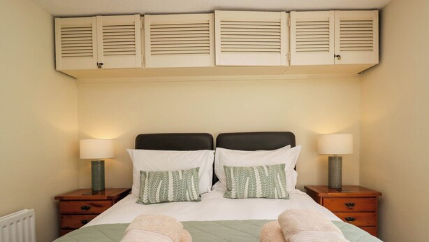Une chambre d’hôtel comprenant un lit, deux tables de chevet, deux lampes, un radiateur et une fenêtre avec des stores.