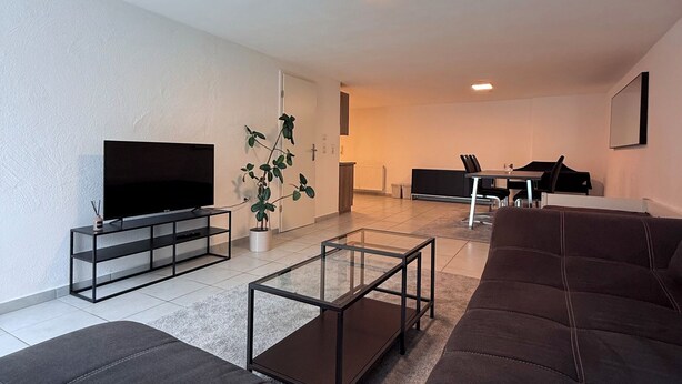 Een moderne woonkamer met een grote flatscreen televisie, een glazen salontafel, een bank en een eethoek met een tafel en stoelen.