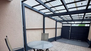 Een overdekt terrasgedeelte met een ronde tafel en twee stoelen, een constructie met metalen frame en een zwart hek op de achtergrond.