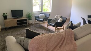 Una sala de estar con un sofá, dos sillones, un televisor sobre un mueble bajo, una ventana con persianas y una planta en maceta.
