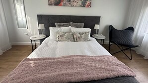 Un dormitorio con una cama grande, dos mesitas de noche, una silla, un cuadro en la pared y una ventana.