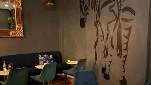 Interior de un restaurante con un sofá de color oscuro, sillas verdes, suelo de madera y un mural con estampados de cebra en la pared.