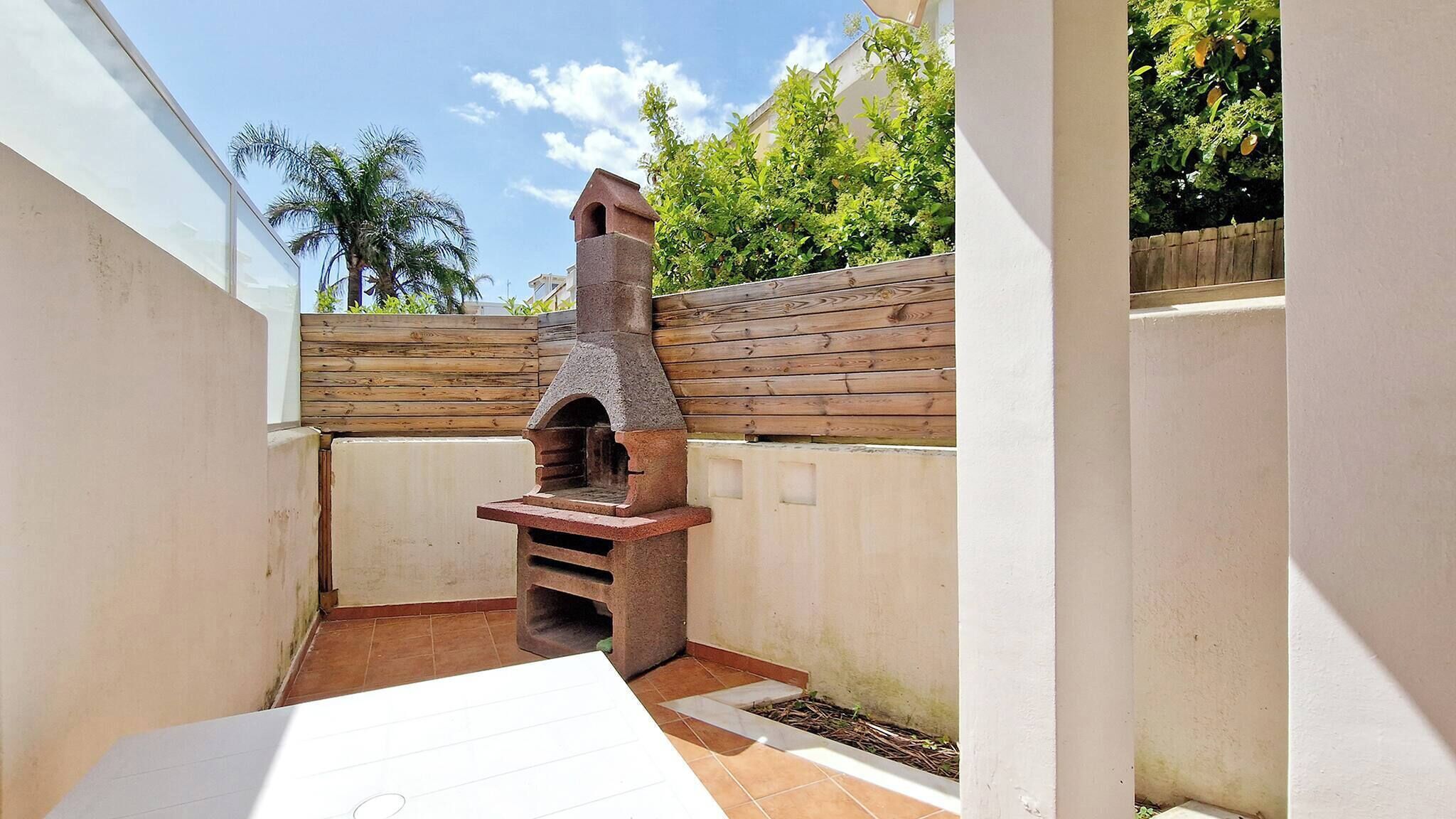Un piccolo patio esterno con un forno in pietra, un recinto in legno e un muro bianco.