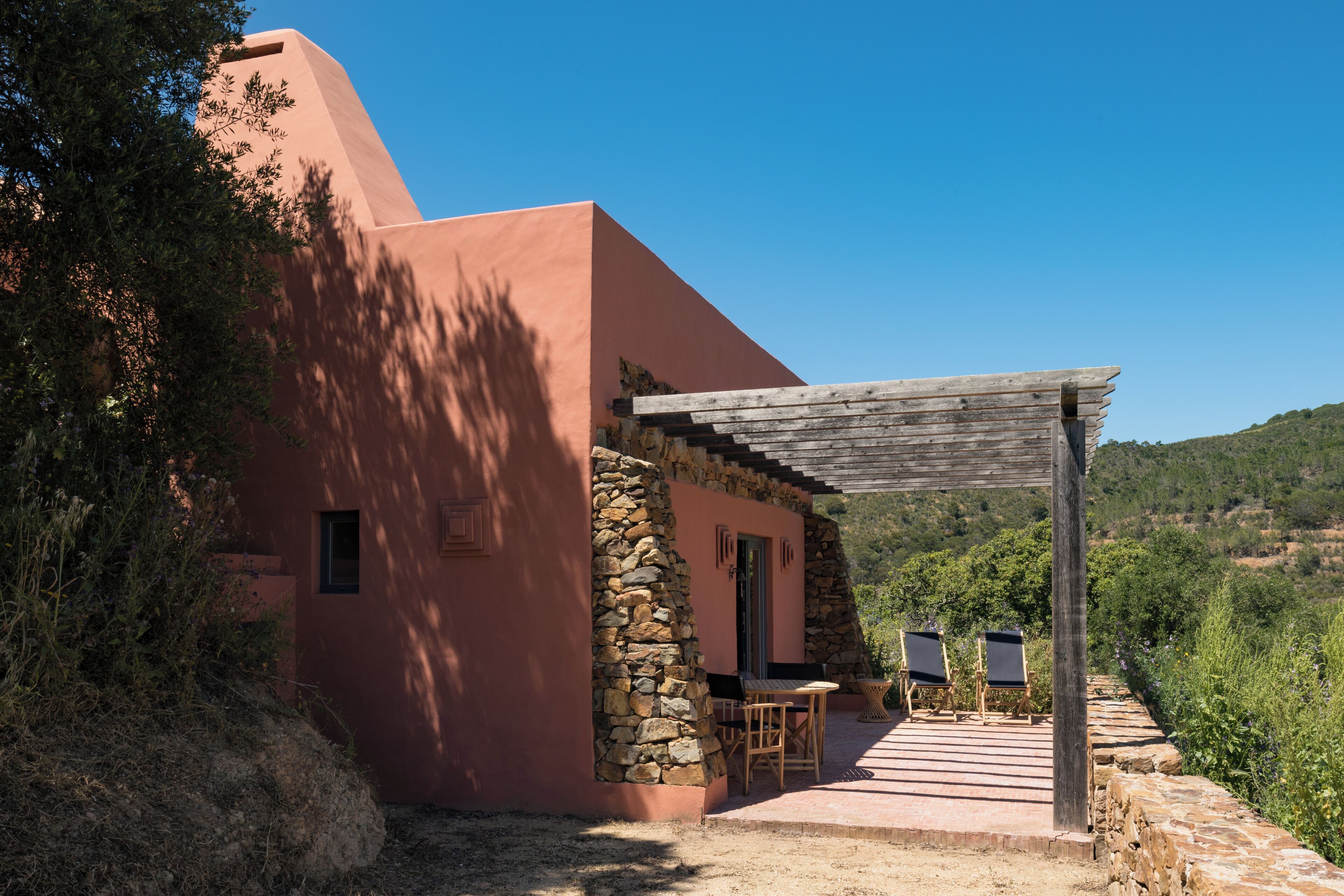 Un edificio rosa con un muro de piedra, una pérgola de madera y dos sillas afuera.
