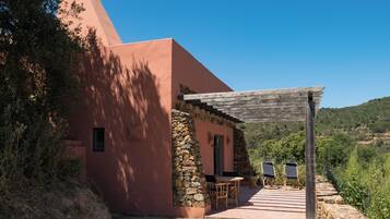 Un edificio rosa con un muro de piedra, una pérgola de madera y dos sillas afuera.