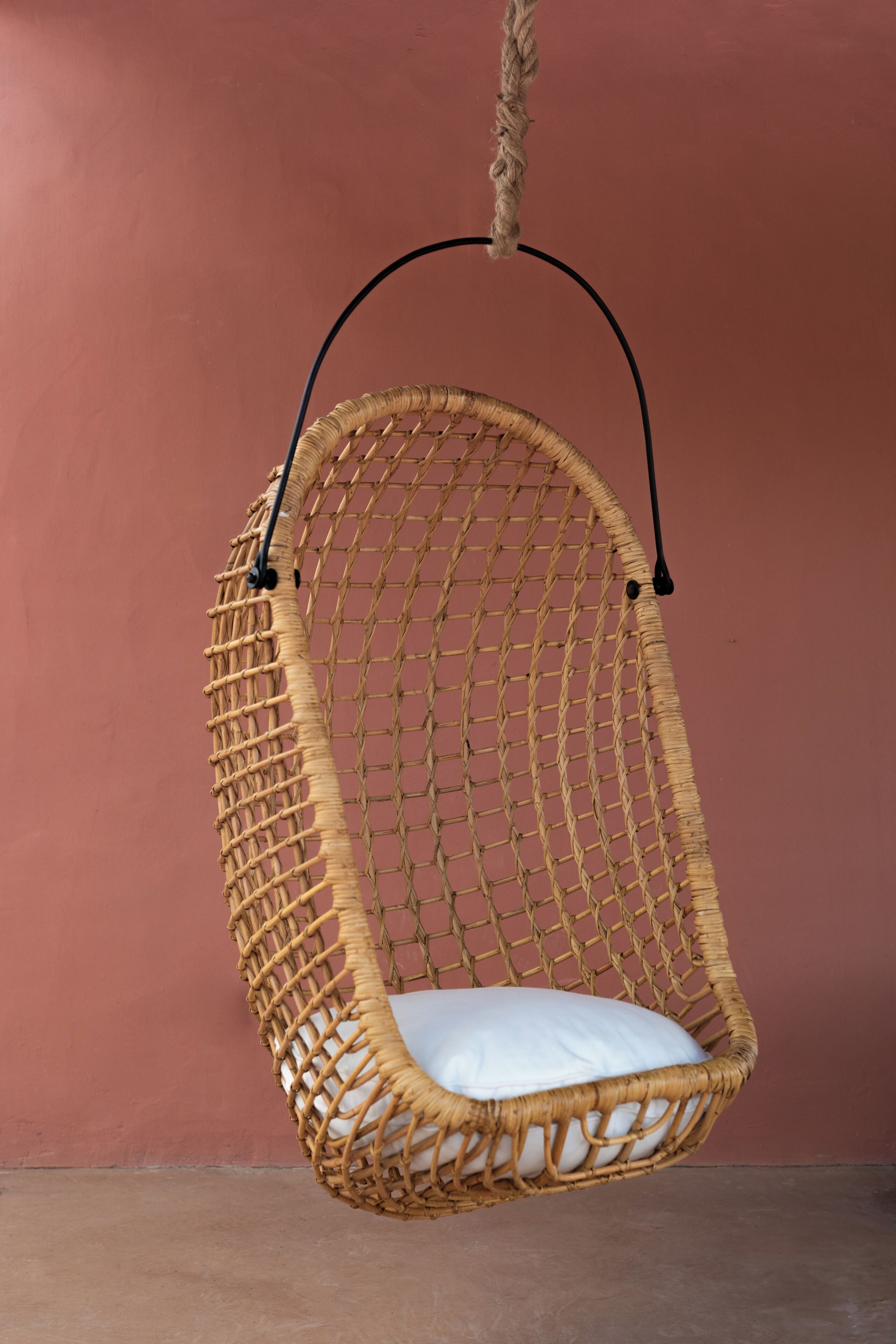 A wicker hanging chair with a white cushion, suspended by a rope against a pink wall.