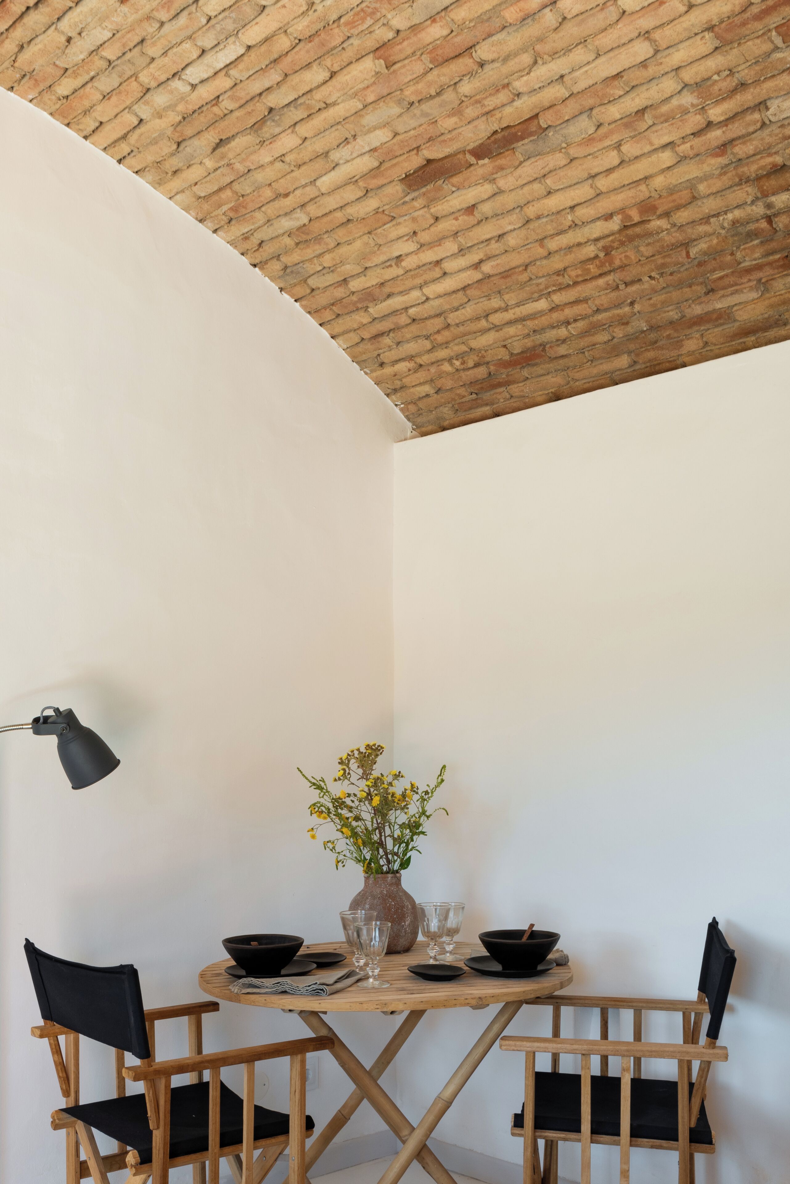 Une pièce d’angle avec un plafond en briques, des murs blancs, une table ronde en bois avec deux chaises, un vase avec des fleurs et un lampadaire noir.
