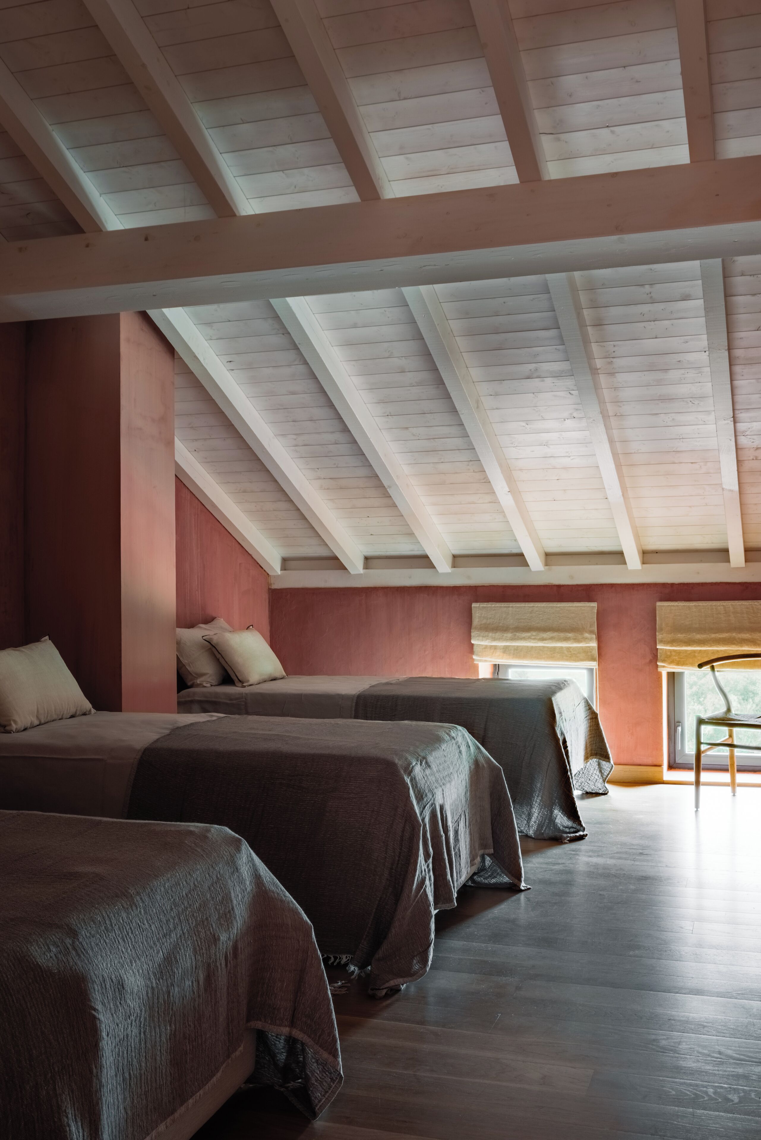A room with three beds aligned against a wall, a large window with blinds, and a sloped ceiling with exposed beams.