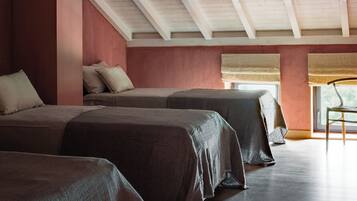 A room with three beds aligned against a wall, a large window with blinds, and a sloped ceiling with exposed beams.