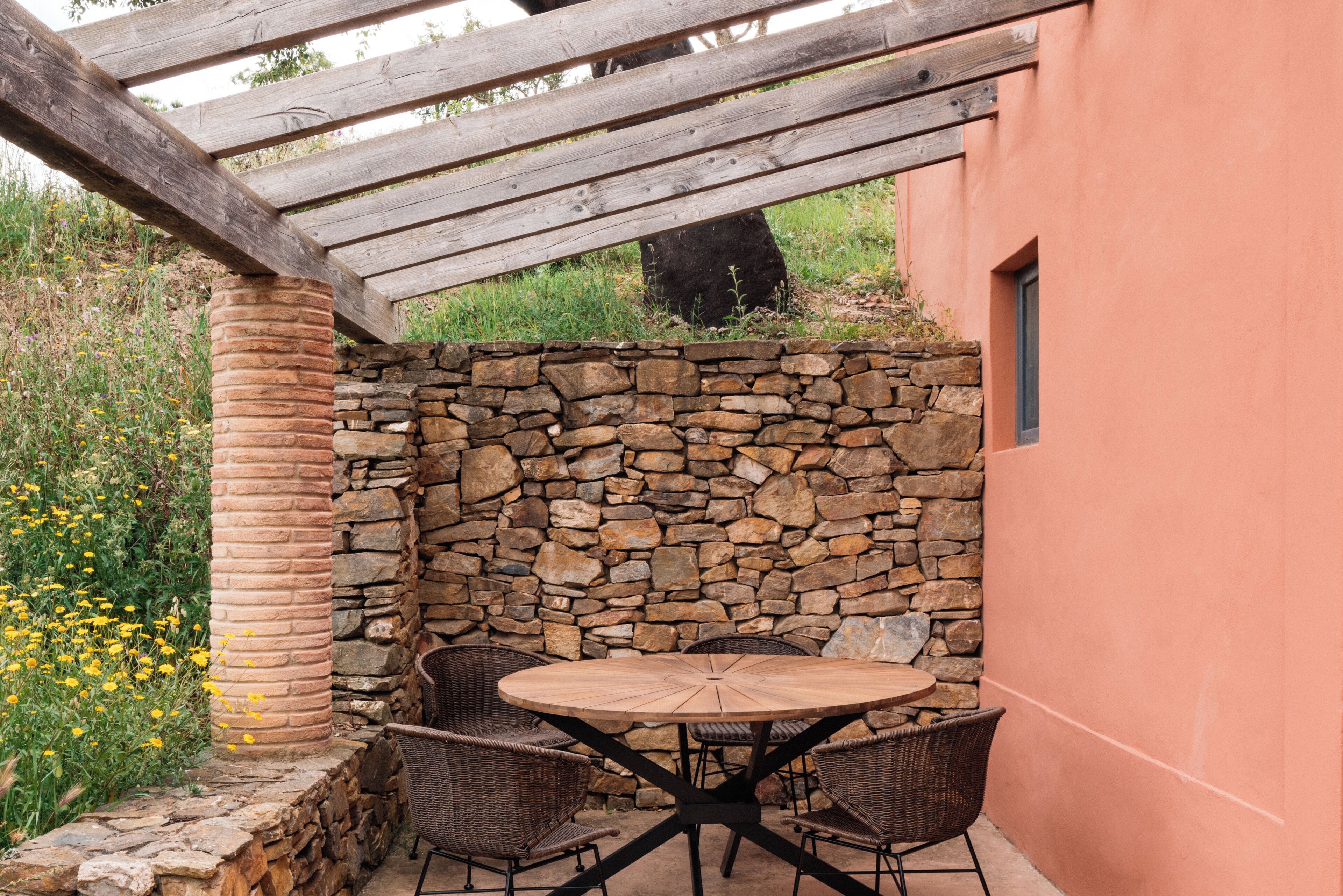 A patio with a stone wall, wooden pergola, and wicker chairs around a round table.