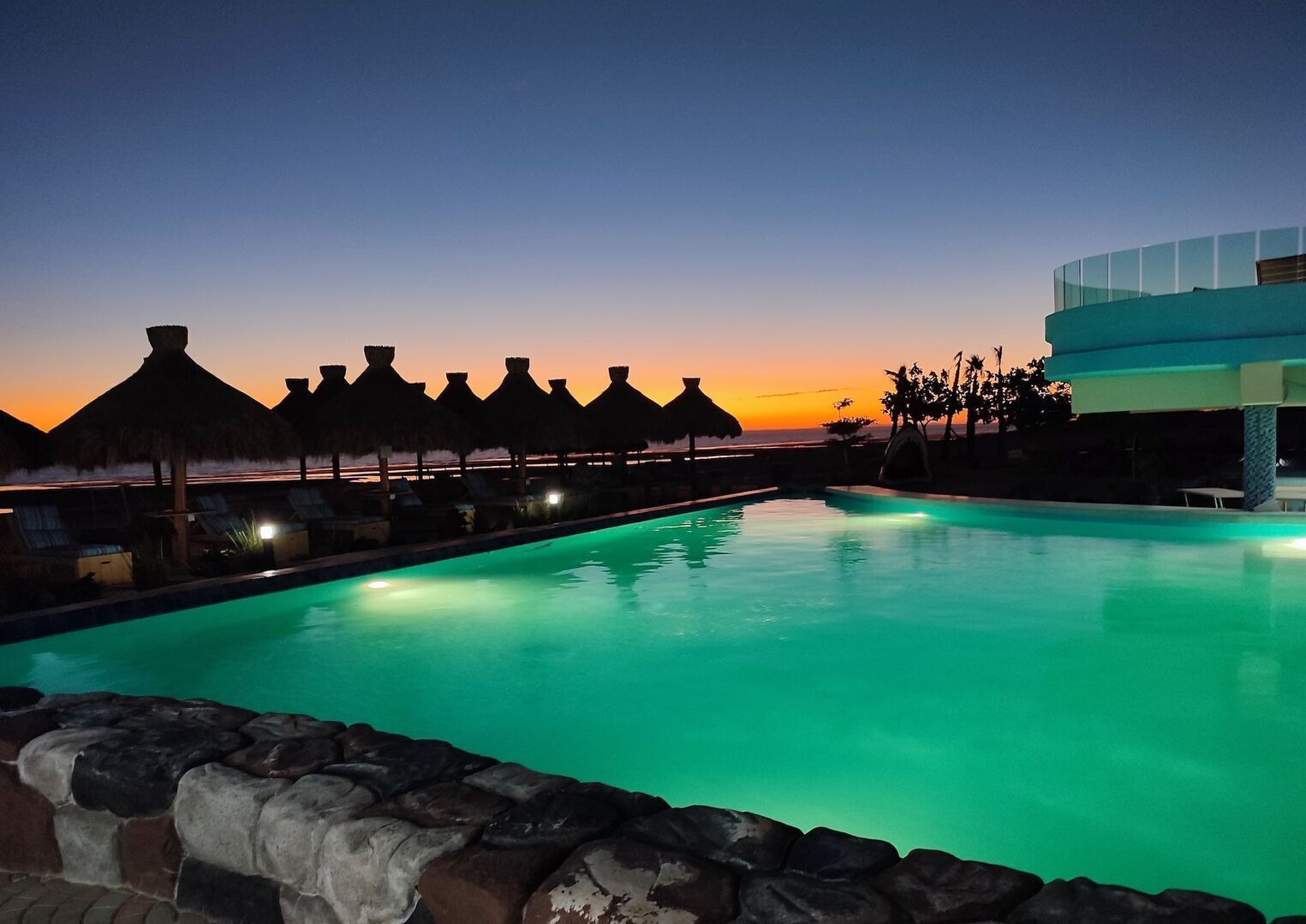 A swimming pool with a sunset view, lounge chairs, and thatched umbrellas.