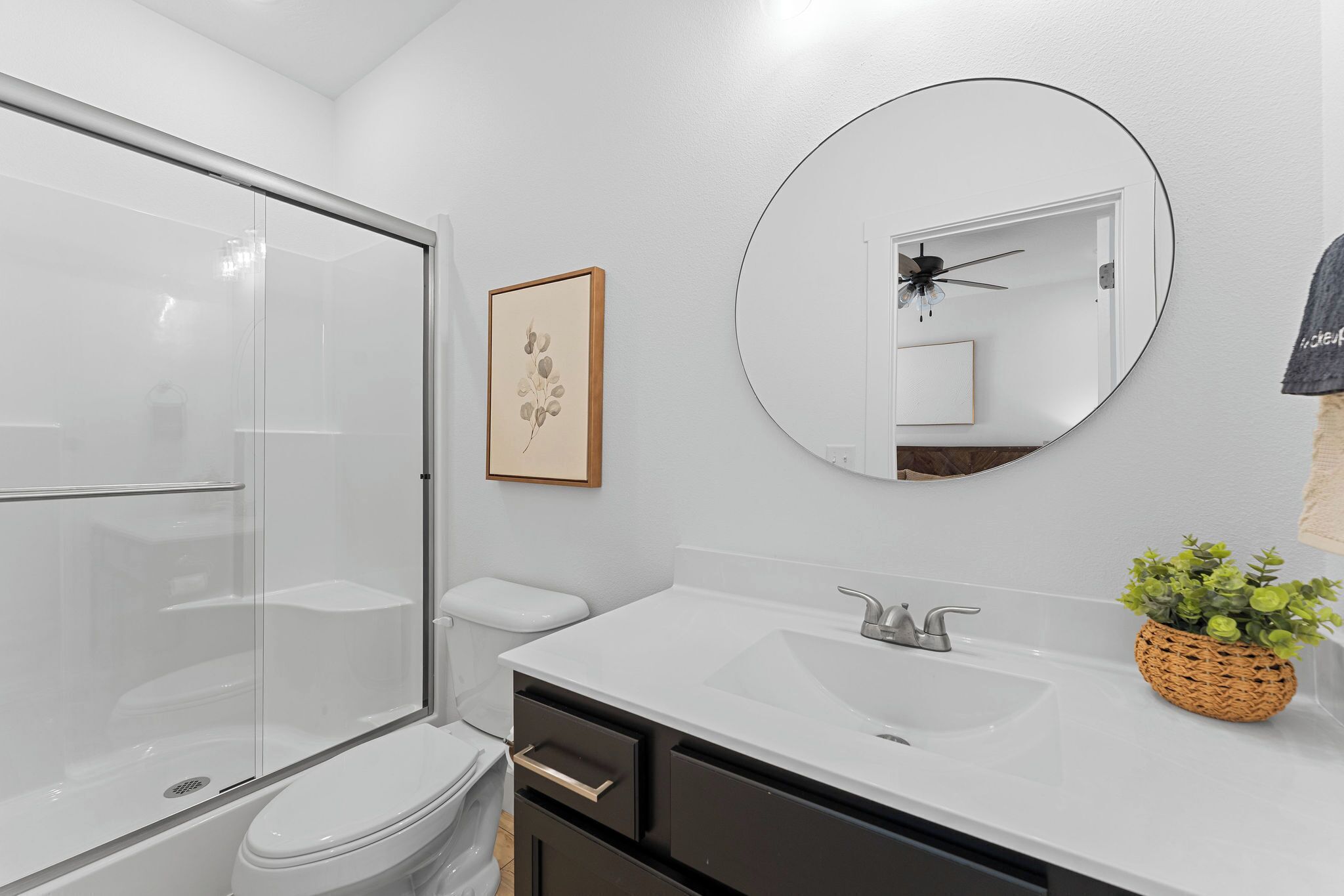 A modern bathroom with a glass shower enclosure, a white sink, a round mirror, and a framed artwork on the wall.