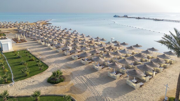 Un complexe hôtelier de bord de mer avec des rangées de chaises longues et de parasols, une jetée qui s’avance dans l’eau et un ciel dégagé.