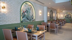 Un restaurant aux murs de briques, aux panneaux de bois vert et aux fresques représentant des scènes tropicales. Les tables sont dressées avec des assiettes, des couverts et des verres.