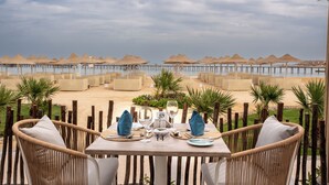 Une table à manger, portant le numéro 14, est entourée de chaises en osier et offre une vue sur une plage parsemée de parasols et sur une jetée.