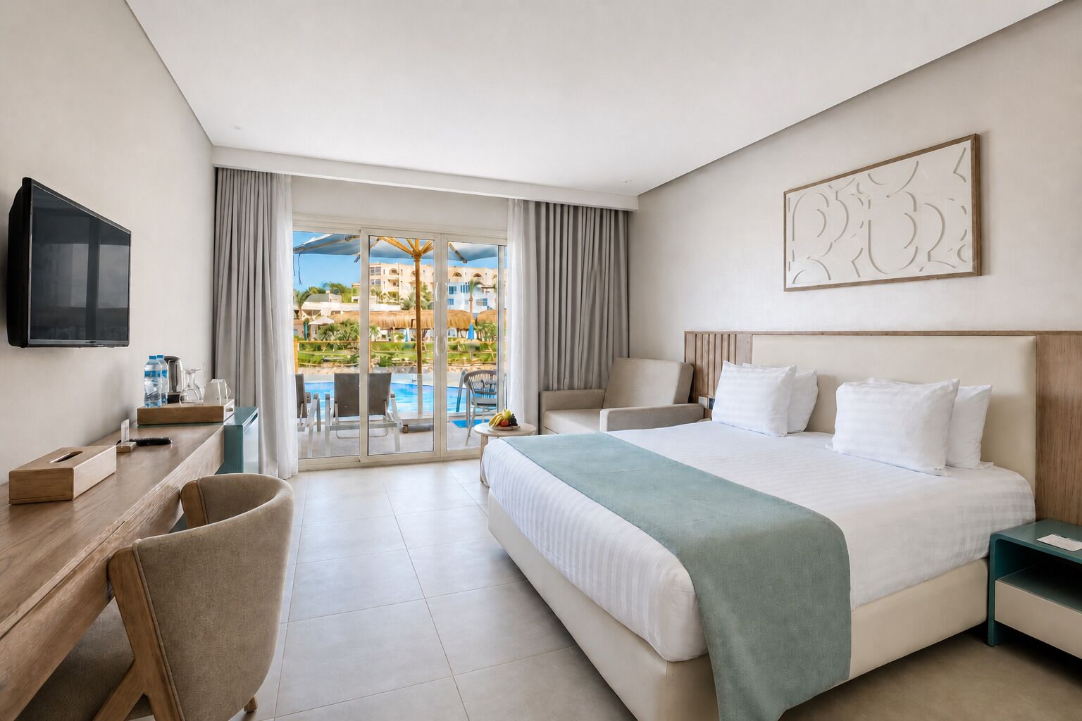 A hotel room with a bed, a desk, a chair, a TV, and a view of a pool and buildings.