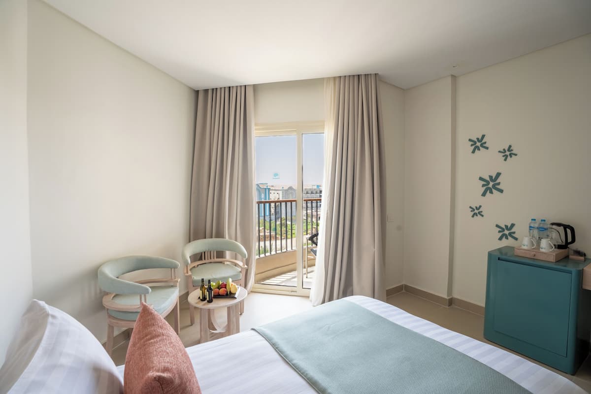 Habitación de hotel con cama, dos sillas, una mesita con botellas, un armario verde azulado y un balcón con cortinas.